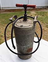 Front view of vintage galvanized metal hand pump fire extinguisher can with wooden handle and black rubber hose coiled around it.
