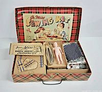 Opened plaid box showing all kit contents arranged inside including doll, fabric, tape measure, patterns and sewing tools.