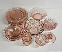 Lot of pink Depression era glassware with diverse glass pieces such as plates, bowls, glasses, and candle holder shown together on table.
