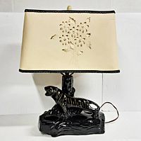 Front view of the vintage black and gold pottery lamp with the cream pierced floral lamp shade installed, showing the cat/tiger figure and floral pattern on the shade.