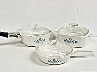 Three vintage Pyrex Corningware Cornflower Blue dishes arranged side by side; includes square casserole, square pan with black handle, and round pan with white handle, all with clear glass lids.