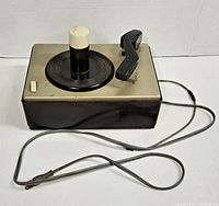 Top and front angle of the vintage RCA Victor turntable showing the black tonearm, white spindle with a protective cap, and the white toggle switch on the silver metal top with brown Bakelite plastic casing.