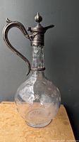 Front view of the antique claret jug showing silvered metal handle, spout and hinged lid with etched glass body featuring star patterns.