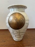 Front view of white ceramic vase with bronze circular geometric design and crackled glaze pattern on wooden surface.