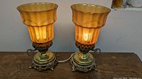 Pair of decorative footed brass table lamps with glowing amber tinted Bohemian art glass shades on wooden surface.