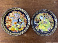 Pair of two Rosenthal artist signed Bjorn Wiinblad Aladin Fantasy ceramic wall plates on wood surface. Both plates have detailed colorful imagery and gold rim designs.