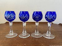 Four cobalt blue etched cordial glasses shown standing in a row on a wooden surface against a plain wall background. The glasses have rounded bowls, clear stems, and flat bases.