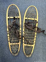 Pair of vintage wooden snow shoes with woven netted design and brown adjustable straps, laid side by side on a blue carpeted surface.