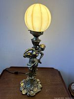 Tall decorative lamp with gold finished lady base and lit art glass ball shade on dark wooden surface against off white wall.