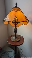 Full view of the vintage stained glass table lamp on a wooden pedestal table showing the amber stained glass shade and bronze base.