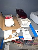 Photo showing the white convertible AMT toy car with red interior positioned beside its original box, along with documentation and additional boxes visible in background.