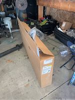 Photo of a cardboard box containing the bumper part in a garage setting with various automotive tools and parts around.