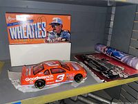 Side view of the die-cast Dale Earnhardt Wheaties stock car on a shelf with branded box behind it and other photo memorabilia to the side.