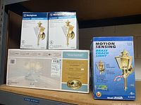 Photo showing boxed Hampton Bay Landmark Plus ceiling fan, two Westinghouse outdoor wall lantern boxes, and boxed Heath/Zenith motion sensing brass light.