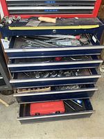 Photo of a five-drawer navy blue tool chest showing some tools inside the partially opened top drawers including wrenches, sockets, and a wooden-handled sharpening tool on top.