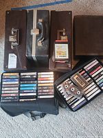 Photo of three closed storage cases and two open storage cases lying on carpet, showing multiple cassette tapes inside. Tapes include various rock and pop artists.