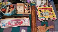 Photo showing a variety of vintage toys including a Barnsbury Puppet box, doll with wigs, toy china tea set box, wooden paddle, and a backgammon game.