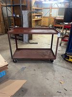 Full view of metal work cart showing two-tier shelving, metal frame, and casters in a workshop setting.