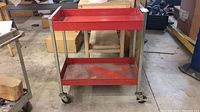 Front view of the red metal work cart showing two shelves and four locking caster wheels on concrete floor in workshop setting.