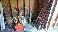 Wide view of multiple yard and garden tools stored inside a wooden shed showing shovels, rakes, wooden stakes, and a post hole digger among others