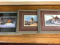 Three framed art prints by Brian Croft arranged side by side on a wooden surface showing detailed scenes including nighttime snow, seaplane, and railway.