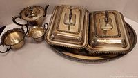 Oval silver plated tray, two lidded rectangular serving dishes, silver plated sugar bowl and creamer (handles visible) and a teapot with hinged lid in the background, all arranged on a plain surface.