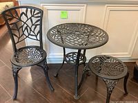 Full view of the metal bistro table and two chairs, showing detailed leaf and web pattern design on all pieces.
