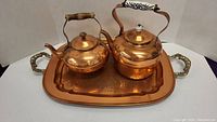 Two polished copper kettles placed on a large copper tray showing wood and ceramic handle details.