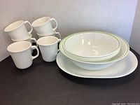 Photo showing three medium bowls, one large bowl, one large platter, and six white mugs organized together.