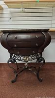 Front view of the end table showing the brown marble top, curved wooden drawers with metal knobs, and ornate black metal base with floral accent.