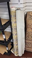 Two bolts of fabric standing upright next to a rug and shelf; one bolt is white vinyl and the other is plaid upholstery fabric.