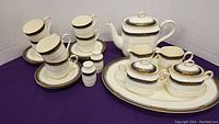 Full tea set including cups stacked on saucers, teapot, platter, salt and pepper, cream and sugar sets on purple surface.