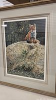 Framed artwork depicting a fox sitting on a rock with plants in the background. The artwork is matted and framed in gold-tone material.