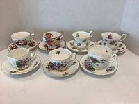 Full set of 7 teacups and 7 saucers displayed in two rows showing floral patterns and gold trim details on porcelain