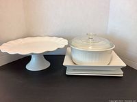 Two square plates stacked with white oval casserole dish with glass lid on top and white pedestal cake stand next to them