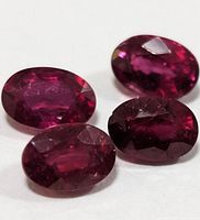 Four oval faceted enhanced ruby gemstones with rich reddish-pink color and visible internal inclusions laid on a white surface.