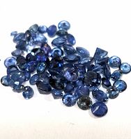 Close-up photo of numerous small blue sapphire gemstones scattered on a flat surface, showing various cuts and sizes.