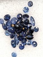 Loose blue sapphire gemstones scattered on light background, various shapes including round and oval cuts, natural inclusions visible