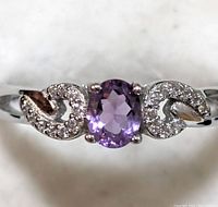 Close-up front view of the ring showing the oval amethyst center flanked by cubic zirconia-studded heart shapes on a sterling silver band.