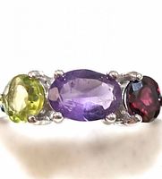 Close up front angle of sterling silver ring with three oval faceted peridot, amethyst, and garnet stones in prong settings.