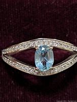 Close-up of ring showcasing oval blue topaz center stone and surrounding cubic zirconia on sterling silver band against dark burgundy background.