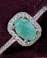 Close-up of sterling silver ring with oval cut green emerald center stone surrounded by clear cubic zirconia stones on the band and halo, on a dark red fabric background.