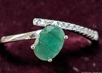 Close-up of the ring showing the oval faceted green emerald held by four prongs and the adjacent band set with small round clear cubic zirconia stones.