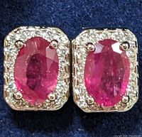 Close-up front view of a pair of sterling silver earrings with oval faceted ruby center stones surrounded by clear cubic zirconia stones.