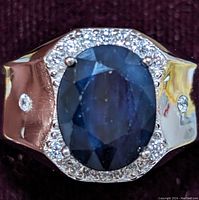 Close-up image showcasing the oval blue sapphire center stone surrounded by clear cubic zirconia on a reflective sterling silver band.