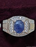Front view of sterling silver men's ring showing oval blue sapphire and surrounding cubic zirconia stones on velvet background