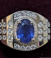Close-up front view of men's ring showing oval blue sapphire centerpiece with round cubic zirconia stones around it and on band sides.