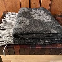 Folded charcoal grey and pale grey leaf patterned merino wool blanket with pale grey fringe on the end.