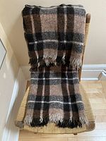 The blanket is folded and draped over a chair, showcasing its plaid pattern in brown, black, cream, and beige stripes with fringed ends.