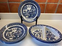 Four stacked Blue Willow soup bowls with one standing behind and one oval divided serving plate in front